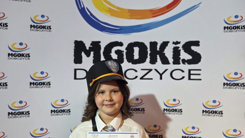uczennica na tle baneru reklamowego MGOKiS Dobczyce. Uczennice trzyma w rękach dyplom. Na głowie ma czapkę służbową dawnego listonosza.