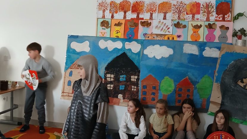 na środku stoi rycerz z mieczem, gotowy do walki ze smokiem Bazyliszkiem. Od lewej strony stoi Szewczyk, który zmierzy się z Bazyliszkiem i znajdzie na niego sposób.