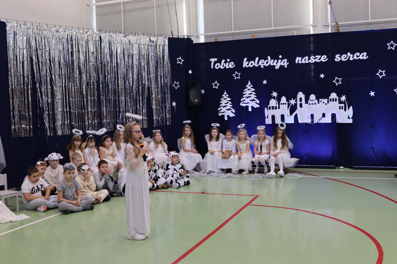 dzieci w strojach aniołów i pastuszków podczas szkolnych jasełek; dziewczynka stoi na środku z mikrofonem, za nią siedzą pozostali uczestnicy na tle świątecznej dekoracji.