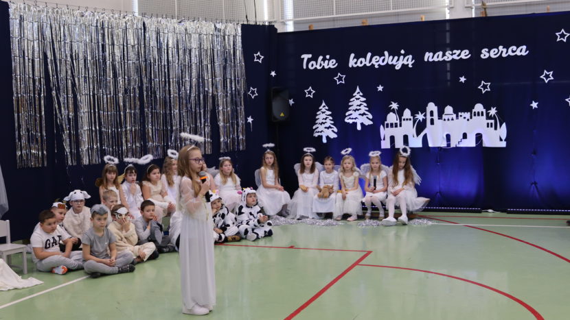 dzieci w strojach aniołów i pastuszków podczas szkolnych jasełek; dziewczynka stoi na środku z mikrofonem, za nią siedzą pozostali uczestnicy na tle świątecznej dekoracji.