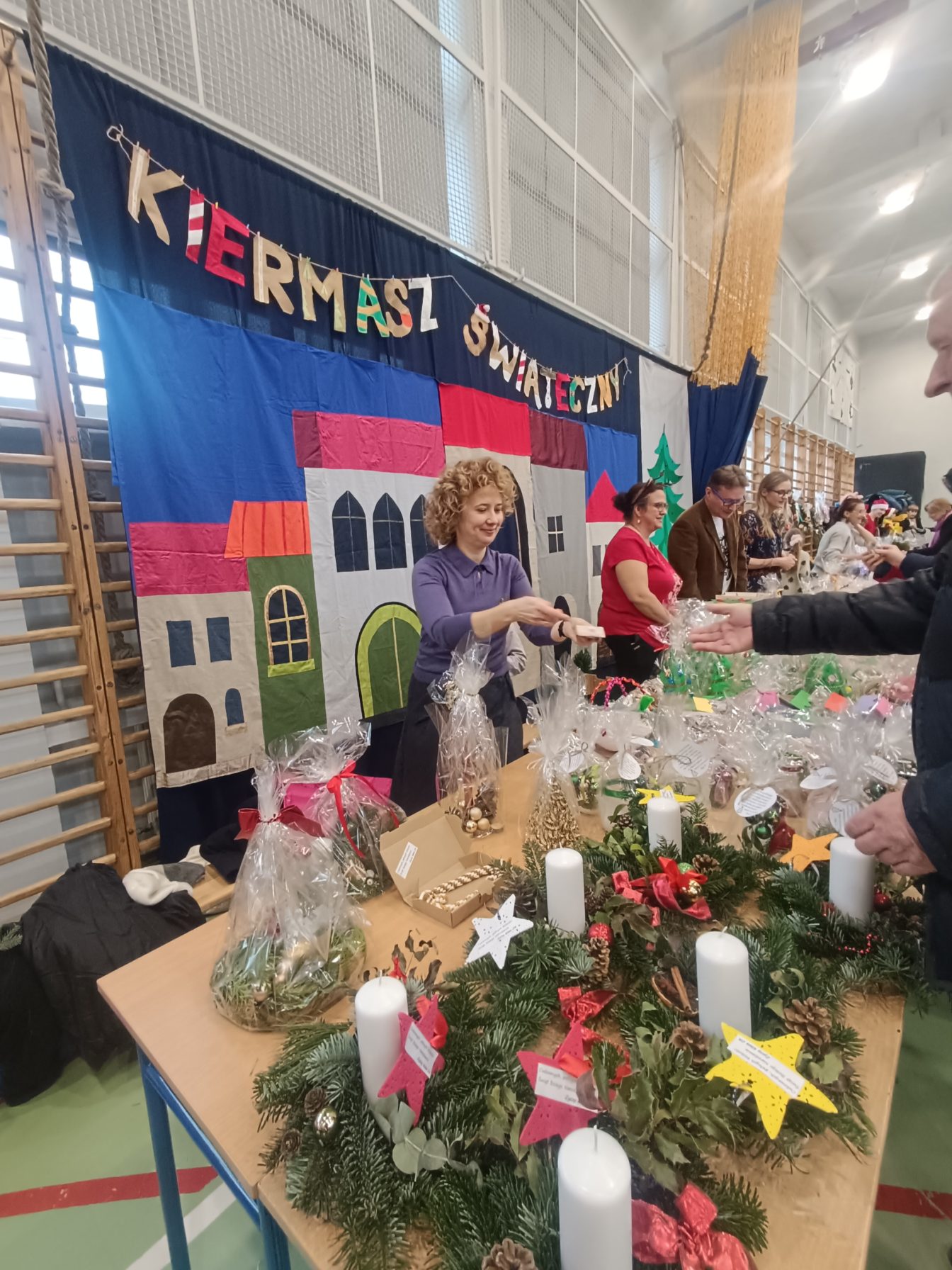 widok sali gimnastycznej SP4 Wieliczka z kiermaszem . W tle grantowe płótno z napisem KIERMASZ ŚWIĄTECZNY