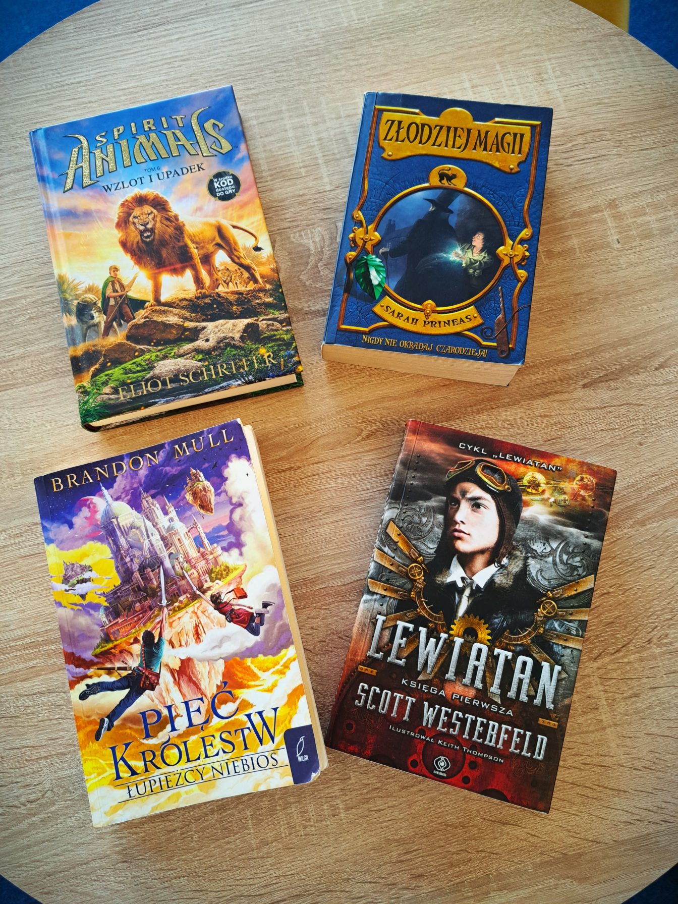 Na okrągłym stole bibliotecznym leżą książki :

1. Zdjęcie z okładkami książek : „Spirit Animals”- na skale stoi lew, a obok niego chłopiec z włócznią i wilk,  „Pięć Królestw”- budynek katedry w stylu gotyku i baroku unoszący się na skale, obok unoszące sie dwie postacie z mieczami w ręku, Złodziej magii”- granatowa okładka, złote litery, w złotym kole postać czarodzieja i chłopca,
, „Lewiatan” - młody mężczyzna w czapce pilotce z okularami na czole na szaro-brązowym tle ....
