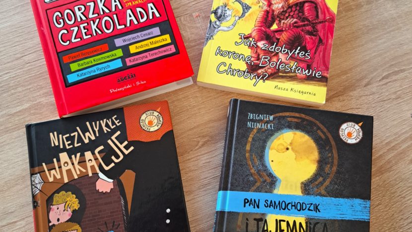 Na okrągłym stole bibliotecznym leżą książki : 1. Zdjęcie z okładkami książek : „Spirit Animals”- na skale stoi lew, a obok niego chłopiec z włócznią i wilk, „Pięć Królestw”- budynek katedry w stylu gotyku i baroku unoszący się na skale, obok unoszące sie dwie postacie z mieczami w ręku, Złodziej magii”- granatowa okładka, złote litery, w złotym kole postać czarodzieja i chłopca, , „Lewiatan” - młody mężczyzna w czapce pilotce z okularami na czole na szaro-brązowym tle .... 2. Zdjęcie z okładkami książek ; „Eliksir przygód”- brązowo –granatowa okładka z rysunkiem zielonego eliksiru oraz głową tajemniczej postaci i dziewczynki z warkoczami, „ BezSennik”- zdjęcie 4 nastolatek ubranych w białe lub czerwonesukienki trzymające czerwonebalony w kształce serc, „ Czekolada z Chili”- jasno beżowe tło ,a na nim niebieski dzbanek z niebieską filiżanką , „ Emi i tajny klub superdziewczyn”- różowa okładka ,po lewo duża postać Emi a obok mniejsze postacie czterech dziewczynek, „ M jak dżem”- okładka w ciepłych kolorach różu i pomarańczu, dziewczynka czyta książkę , w tle regały z przetworami. 3. Zdjęcie z okładkami książek : „Gorzka czekolada”- okładka w kolorze czerwonym u góry widoczne nogi dzieci, „ Jak zdobyłeś koronę Bolesławie Chrobry” -żółta okładka w środku siedzi zamyślony król, „ Niezwykłe wakacje”- dwoje dzieci patrzących na umykającą postać, „ Pan Samochodzik i tajemnica tajemnic”- czarna okładka z żółtą dziurką od klucza.