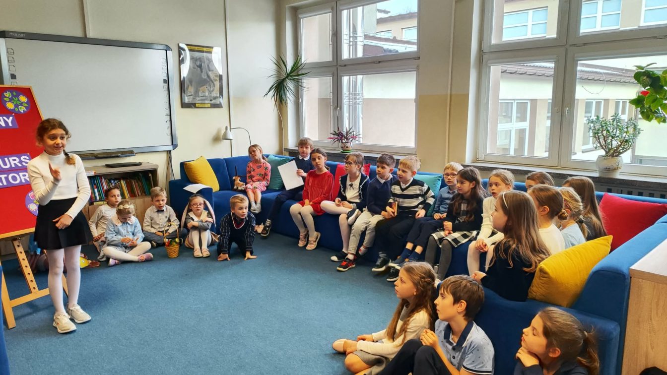 Uczennica recytuje przed publicznoscią zebraną w bibliotece szkolnej