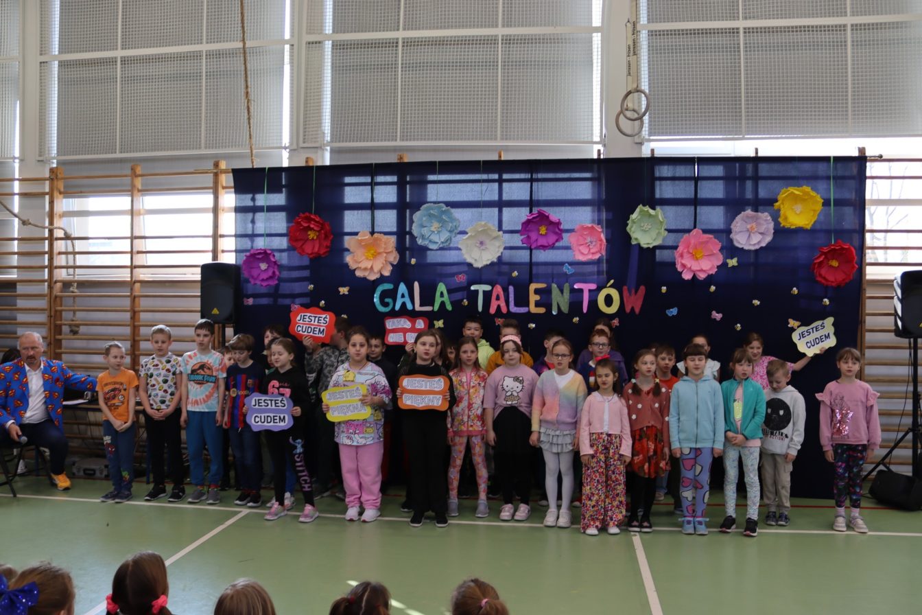 Dzieci uczestniczące w Gali Talentów w SP4 Wieliczka stoją w sali gimnatycznej, w tle dekoracja na granatowym płótnie  napisem Gala Talentóa