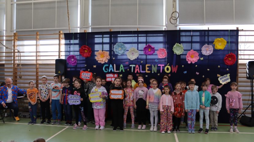 Dzieci uczestniczące w Gali Talentów w SP4 Wieliczka stoją w sali gimnatycznej, w tle dekoracja na granatowym płótnie napisem Gala Talentóa