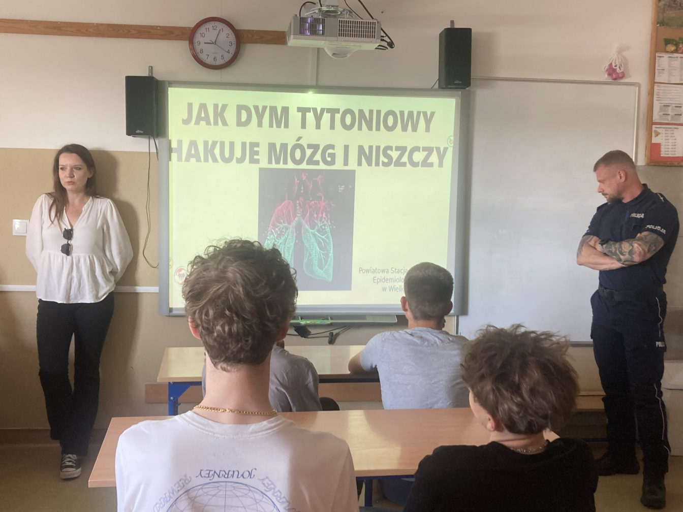 Sala lekcyjna podczas spotkania profilaktycznego dotyczącego szkodliwości palenia tytoniu. Przed uczniami stoi prelegentka oraz funkcjonariusz policji. Na ekranie wyświetlona jest prezentacja zatytułowana „Jak dym tytoniowy hakuje mózg i niszczy”, ilustrowana grafiką płuc.”