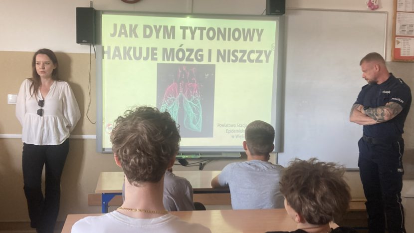 Sala lekcyjna podczas spotkania profilaktycznego dotyczącego szkodliwości palenia tytoniu. Przed uczniami stoi prelegentka oraz funkcjonariusz policji. Na ekranie wyświetlona jest prezentacja zatytułowana „Jak dym tytoniowy hakuje mózg i niszczy”, ilustrowana grafiką płuc.”