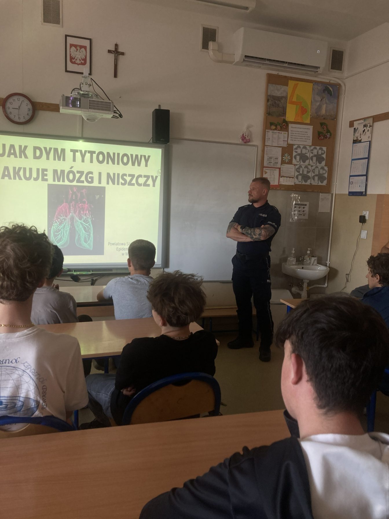 Uczniowie siedzą w klasie i słuchają prelekcji na temat wpływu dymu tytoniowego na zdrowie. Obok ekranu z prezentacją stoi funkcjonariusz policji.