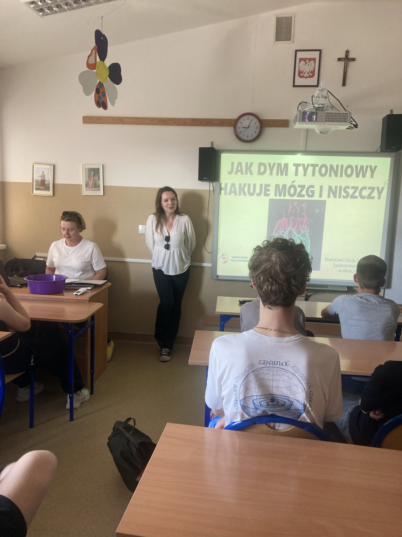 Szkolne zajęcia profilaktyczne poświęcone zagrożeniom związanym z paleniem tytoniu. Przed klasą stoją dwie prowadzące, a na ekranie wyświetlona jest prezentacja edukacyjna dotycząca wpływu dymu tytoniowego na mózg i organizm. Uczniowie słuchają wykładu z ławek.